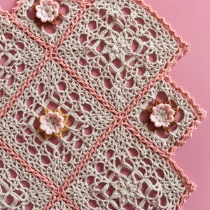 Può includere: Una coperta all'uncinetto con sfondo beige e dettagli rosa e marroni. La coperta è realizzata con un delicato motivo a pizzo e presenta un bordo a festoni.