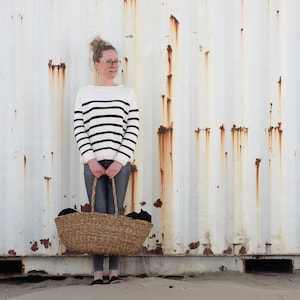 May include: A woman wearing a white and black striped sweater and blue jeans is holding a woven basket with black fabric inside. The woman is standing in front of a white metal wall with rust colored streaks.