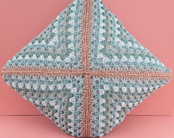 May include: A square, crocheted pillow in shades of blue, gray, and white, with a cross-shaped, light brown border. The pillow has a textured, open weave pattern, and is set against a pink background.