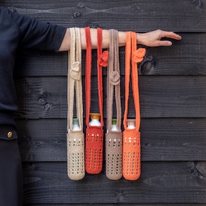 Puede incluir: Cuatro porta botellas de ganchillo en naranja, beige y verde colgados de una pared de madera. Los porta botellas están hechos con un patrón de punto simple y tienen una correa para llevar.