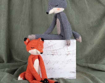 May include: A gray crocheted wolf and an orange crocheted fox sitting on a white wooden box. The wolf is sitting with its legs crossed and its head tilted to the side. The fox is sitting with its legs stretched out in front of it.