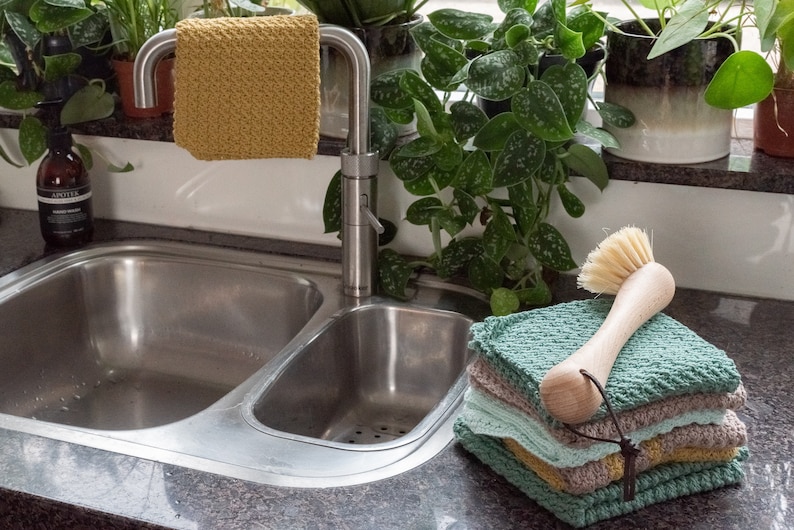 May include: A stainless steel kitchen sink with two basins. A yellow dishcloth hangs on the faucet. A stack of green and brown dishcloths sits on the counter next to a wooden dish brush.