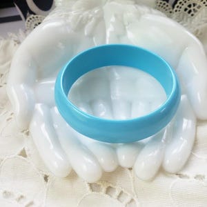 May include: A smooth, solid light blue bangle bracelet with a rounded exterior surface. The bracelet is displayed resting on a white, opaque glass sculpture shaped like two cupped hands, which is set upon a white lace fabric.
