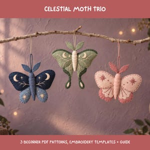 Puede incluir: Tres adornos colgantes de fieltro con forma de polilla etiquetados como CELESTIAL MOTH TRIO. La polilla izquierda es azul oscuro con bordados blancos de lunas y estrellas. La central es verde salvia con detalles lunares. La derecha es rosa claro.