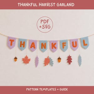 May include: A Thankful Harvest Garland banner featuring light purple and blue pennants with orange letters spelling out THANKFUL. Below the pennants hang various felt shapes including orange maple leaves, acorns, and blue and purple leaves.