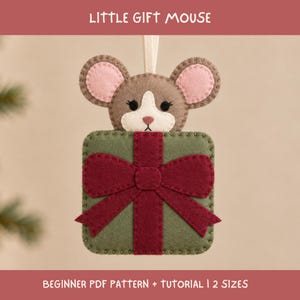 Può includere: Un ornamento in feltro a forma di testa di topo che spunta da un pacchetto regalo quadrato verde con un fiocco rosso scuro. Il topo ha il pelo marrone, l'interno delle orecchie rosa e il muso bianco. Il testo LITTLE GIFT MOUSE e BEGINNER PDF PATTERN + TUTORIAL | 2 SIZES è visibile.