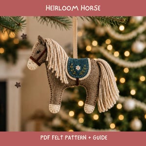 May include: A felt horse ornament with a brown body, cream-colored yarn mane and tail, and a dark teal saddle featuring embroidered floral details. The horse has a brown bridle and is suspended by a beige ribbon. Text reads HEIRLOOM HORSE.
