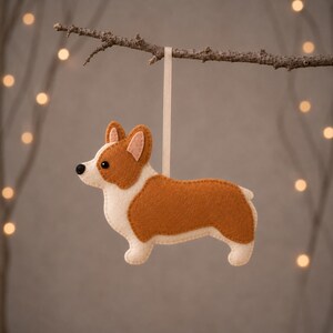 May include: A felt Corgi ornament featuring a tan and cream-colored body with a black nose and eye. The dog-shaped decoration is crafted from layered felt fabric and hangs from a thin, light-colored ribbon loop attached to its back.