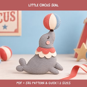Peut inclure: Une décoration en feutrine grise représentant un phoque avec une collerette à volants rouge et crème, équilibrant une balle rouge, crème et bleue sur son nez. Le texte "LITTLE CIRCUS SEAL" et "PDF + SVG PATTERN & GUIDE | 2 SIZES" est visible.