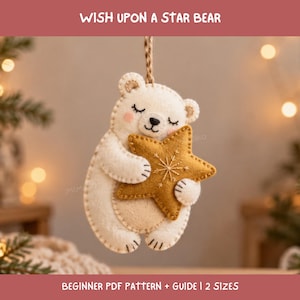 May include: A felt ornament shaped like a white bear with closed eyes and pink cheeks, holding a mustard yellow star with white embroidered snowflake details. The bear has a tan belly patch and is suspended by a brown twine loop for hanging.