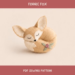 May include: A tan felt fennec fox ornament with large ears, closed eyes, and a black nose. The fox is curled in a sleeping position and features embroidered green leaves and small pink flowers on its side. The text FENNEC FOX and PDF SEWING PATTERN is visible.