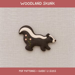 Può includere: Una sagoma in feltro marrone scuro a forma di puzzola con una striscia color crema che va dalla testa alla coda. La puzzola presenta fiori rosa ricamati con steli verdi sul fianco. È visibile il testo WOODLAND SKUNK e PDF PATTERNS + GUIDE | 2 SIZES.