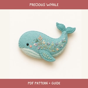May include: A teal felt whale ornament featuring delicate floral embroidery in shades of pink, purple, and white across its side. The whale has a small black bead eye, a pink cheek detail, and a curved tail. Text reads PRECIOUS WHALE PDF PATTERN + GUIDE.