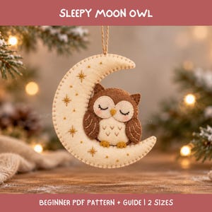 Op de afbeelding: Een vilten ornament in de vorm van een crèmekleurige halve maan met geborduurde gouden sterren en een bruine uil met gesloten ogen. De tekst SLEEPY MOON OWL en BEGINNER PDF PATTERN + GUIDE | 2 SIZES is zichtbaar op de afbeelding.