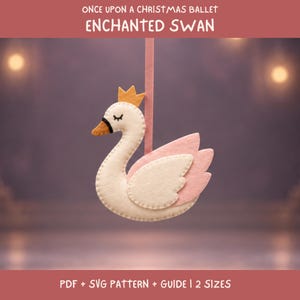 Puede incluir: Un adorno de cisne de fieltro con cuerpo color crema, corona dorada y alas rosa claro. El cisne tiene ojos cerrados bordados y un pico negro y naranja. Cuelga de una cinta rosa. El texto dice: ONCE UPON A CHRISTMAS BALLET ENCHANTED SWAN.