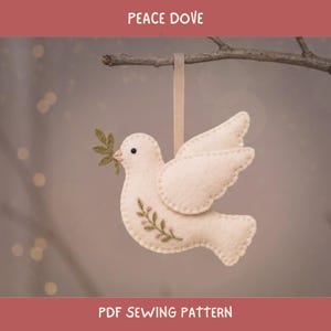 May include: A cream-colored felt dove ornament hanging from a branch by a beige ribbon. The dove features a small black eye, a green felt olive branch in its beak, and an embroidered green vine with tiny pink flowers on its side. Text reads PEACE DOVE.