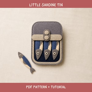 Puede incluir: Un proyecto de manualidades en fieltro titulado LITTLE SARDINE TIN PDF PATTERN + TUTORIAL. La imagen muestra una lata rectangular de fieltro gris que contiene tres formas de peces de fieltro azul y beige, con un pez a juego a la izquierda.