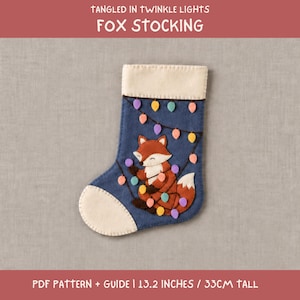 May include: A blue felt Christmas stocking featuring an applique of a fox tangled in colourful string lights. The stocking has a cream-coloured cuff and toe. Text at the top reads TANGLED IN TWINKLE LIGHTS FOX STOCKING and PDF PATTERN + GUIDE 33 CM TALL.