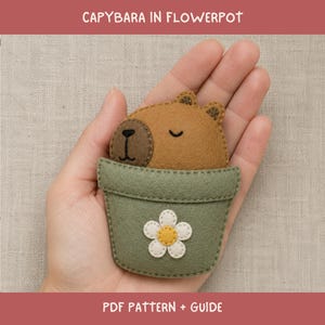 May include: A felt craft project featuring a brown capybara with embroidered facial features tucked inside a sage green flowerpot. The pot is decorated with a white and yellow flower applique. The text reads CAPYBARA IN FLOWERPOT PDF PATTERN + GUIDE.