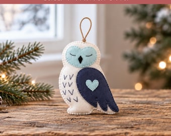 May include: A felt owl ornament featuring a white body with dark blue wings, a light blue face with closed eyes, and a small light blue heart on the wing. The owl has a brown twine loop for hanging and includes the text Heart of Winter Sleepy Winter Owl.