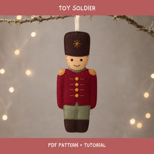 Puede incluir: Un adorno de fieltro de un soldado de juguete con gorro marrón oscuro y estrella amarilla, chaqueta roja con botones amarillos y pantalones verde oliva. La figura tiene cara beige con una sonrisa cosida. Texto: TOY SOLDIER PDF PATTERN + TUTORIAL.