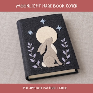 Può includere: Una copertina per libro in feltro grigio scuro con un design ad applicazione che raffigura una luna beige chiaro, una lepre marrone chiaro che guarda verso l'alto e rami frondosi viola chiaro. Tre stelle azzurre sono sopra la luna.