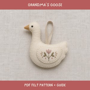 Pode incluir: Um ornamento de feltro cor de creme em forma de ganso com bico bege, um pequeno olho preto e uma fita bege para pendurar. A asa é decorada com detalhes florais bordados em rosa e verde. Texto: GRANDMA'S GOOSE PDF FELT PATTERN + GUIDE.