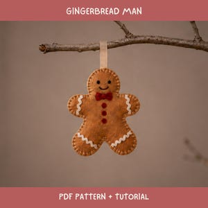 May include: A tan felt gingerbread man ornament featuring white zigzag stitching on the arms and legs, a red bow tie, and three red circular buttons. The figure has a stitched smile and eyes, hanging from a beige ribbon loop. Text reads GINGERBREAD MAN PDF PATTERN + TUTORIAL.
