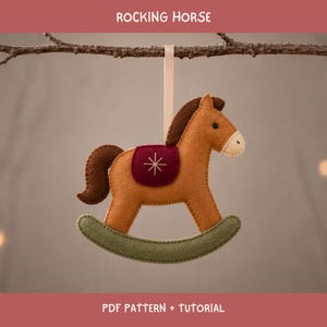 May include: A felt rocking horse ornament with a tan body, dark brown mane and tail, a maroon saddle featuring a white stitched star, and olive green rockers. The ornament hangs from a light beige ribbon and includes the text ROCKING HORSE.