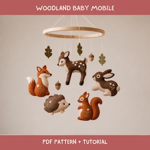 May include: A woodland baby mobile featuring felt figures of a fox, deer, rabbit, hedgehog, and squirrel suspended from a circular wooden frame. The figures are decorated with embroidered white flowers. Includes text: WOODLAND BABY MOBILE PDF PATTERN + TUTORIAL.