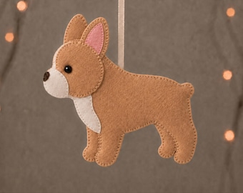 May include: A tan felt French bulldog ornament with a white muzzle and chest, pink inner ears, and a dark brown nose. The ornament hangs from a light-colored ribbon. The text "FRENCH BULLDOG" and "PDF & SVG FELT SEWING PATTERN + GUIDE" is visible.