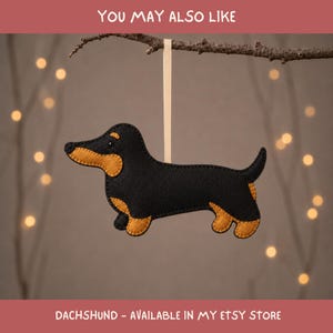 May include: A black and tan felt dachshund ornament featuring a stitched edge and a small black eye. The dog figure is suspended by a thin cream-colored ribbon attached to its back, designed for hanging as a decorative piece.