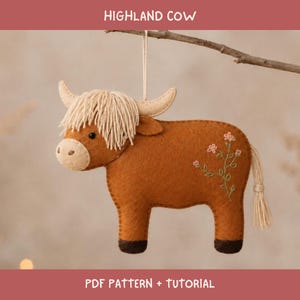 Könnte beinhalten: Ein Highland-Rind-Anhänger aus Filz in Rostbraun mit einer cremefarbenen Garnfransenmähne, hellbeigen Hörnern und dunkelbraunen Hufen. Der Körper ist mit gestickten rosa und grünen Blumendetails sowie einer cremefarbenen Schwanzquaste verziert.