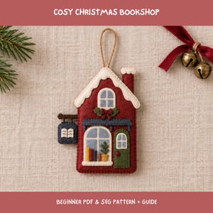 May include: A felt ornament shaped like a house with a red exterior, white snow-covered roof, and a chimney. The front features a green door, a window with colorful books and a plant, and a hanging sign with an open book icon. Includes a string loop.