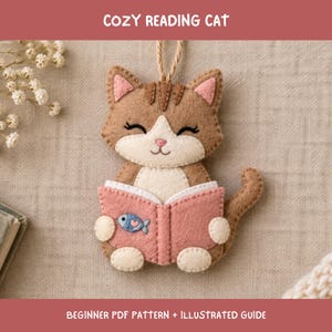 Könnte beinhalten: Ein Filzanhänger in Form einer braun-weißen Katze, die ein rosa Buch mit einem kleinen blauen Fischsymbol auf dem Einband liest. Die Katze hat geschlossene Augen, rosa Innenohren und eine Juteschnur. Der Text COZY READING CAT und BEGINNER PDF PATTERN + ILLUSTRATED GUIDE ist sichtbar.