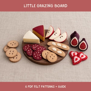 Puede incluir: Un conjunto de alimentos de fieltro sobre una tabla de madera redonda, incluyendo trozos de queso, rodajas de manzana, galletas, higos y corazones. El texto "LITTLE GRAZING BOARD" y "6 PDF FELT PATTERNS + GUIDE" es visible en la imagen.