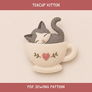 May include: A felt craft project featuring a grey and white kitten peeking out of a cream-colored teacup. The teacup is decorated with a small pink heart and green embroidered sprigs. The text TEACUP KITTEN and PDF SEWING PATTERN is visible.
