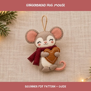 May include: A felt mouse ornament featuring the text GINGERBREAD HUG MOUSE and BEGINNER PDF PATTERN + GUIDE. The mouse is light brown with pink inner ears, a dark red scarf, and holds a brown heart. It has a hanging loop and a pink tail.