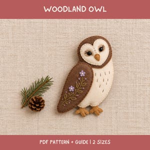 May include: A felt owl craft project featuring the text WOODLAND OWL and PDF PATTERN + GUIDE | 2 SIZES. The owl is brown and cream with embroidered purple flowers on its wing, black bead eyes, and a small pinecone and evergreen sprig nearby.