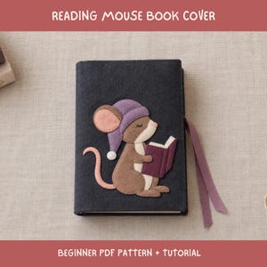 May include: A dark charcoal grey felt book cover featuring an appliqué of a brown mouse wearing a purple nightcap and reading a purple book. The cover includes a purple ribbon bookmark attached to the side, with the text READING MOUSE BOOK COVER.