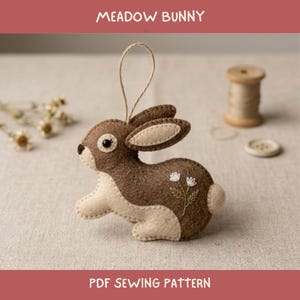 Pode incluir: Um ornamento de coelho de feltro castanho e creme com uma alça para pendurar, apresentando flores brancas bordadas na lateral. O texto MEADOW BUNNY e PDF SEWING PATTERN é exibido. O coelho tem um olho escuro e detalhes costurados nas orelhas.