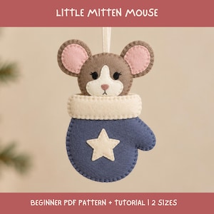 Può includere: Un ornamento in feltro a forma di muffola blu con una stella e un polsino color crema, caratterizzato da una testa di topo marrone con orecchie rosa che spunta dalla parte superiore. Il testo LITTLE MITTEN MOUSE e BEGINNER PDF PATTERN + TUTORIAL | 2 SIZES è visibile.