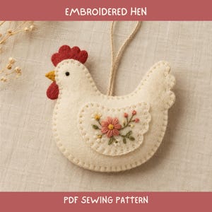 Op de afbeelding: Een crèmekleurig vilt hen-ornament met een rode kam en lellen, een gele snavel en een zwart oog. De vleugel is versierd met geborduurde roze en witte bloemen en groene blaadjes. Een beige touwtje is bevestigd om het ornament op te hangen.