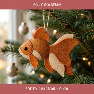 May include: A felt goldfish ornament featuring a layered design in shades of orange and cream with a small black bead eye. The fish is suspended by a thin twine loop and displays the text GILLY GOLDFISH and PDF FELT PATTERN + GUIDE.