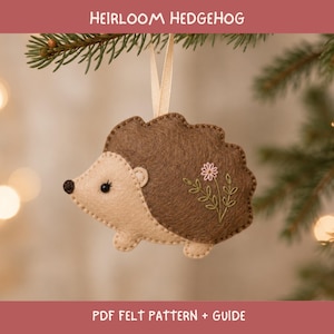 May include: A felt hedgehog ornament featuring a light beige face and a dark brown back with embroidered floral details. The hedgehog has a small black nose and eye, and is suspended by a light beige ribbon. The text HEIRLOOM HEDGEHOG PDF FELT PATTERN + GUIDE is visible.