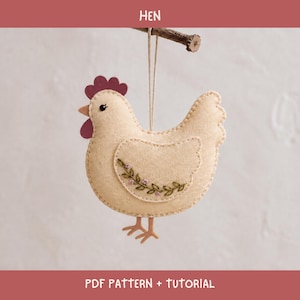 May include: A cream-coloured felt hen ornament hanging from a wooden twig. The hen features a dark red comb and wattle, a small black eye, and an embroidered green vine with tiny pink flowers on its wing. The text HEN PDF PATTERN + TUTORIAL is visible.