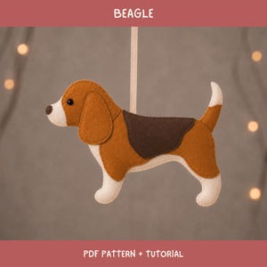 May include: A felt beagle ornament featuring brown, dark brown, and cream-colored sections with a black eye and nose. The dog is suspended by a light-colored ribbon. The text BEAGLE and PDF PATTERN + TUTORIAL are printed on the top and bottom borders.