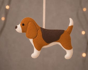 May include: A felt beagle ornament featuring brown, dark brown, and cream-colored sections with a black eye and nose. The dog is suspended by a light-colored ribbon. The text BEAGLE and PDF PATTERN + TUTORIAL are printed on the top and bottom borders.