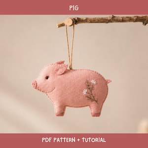 Könnte beinhalten: Ein hellrosa Filzschwein-Ornament, das an einer braunen Holzstange an einer dünnen Schnur hängt. Das Schwein hat ein kleines gesticktes Auge und ein zartes Blumenmuster, das mit weißem und braunem Garn auf die Seite gestickt ist. Der Text PIG PDF PATTERN + TUTORIAL ist sichtbar.