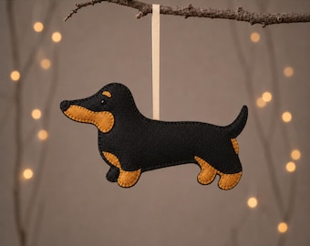 May include: A black and tan felt dachshund ornament hanging from a light-colored ribbon. The dog-shaped decoration features stitched edges and is displayed with the text DACHSHUND at the top and PDF & SVG PATTERN + GUIDE at the bottom.